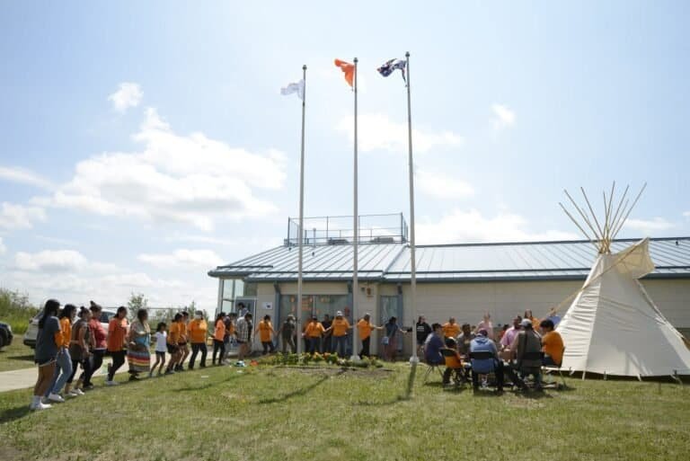 Community members in orange shirts round dancing in front of the Red Pheasant Health Clinic, with drummers beside a tipi