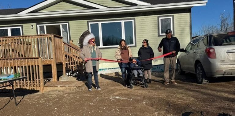 A ribbon cutting in front of a newly built green home, with a leader in a feather headdress and community members holding the red ribbon